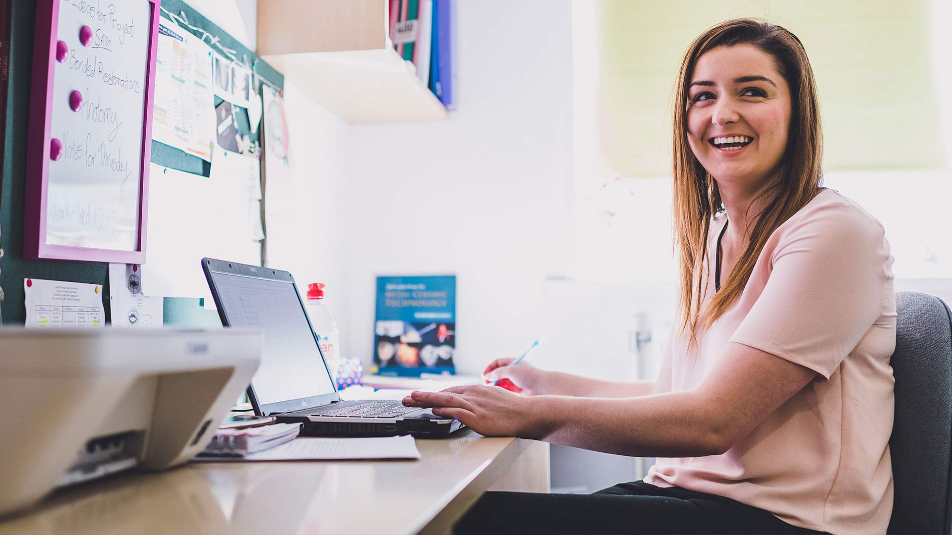 Smiling woman working at her desk with a laptop, taking notes in a bright office space.