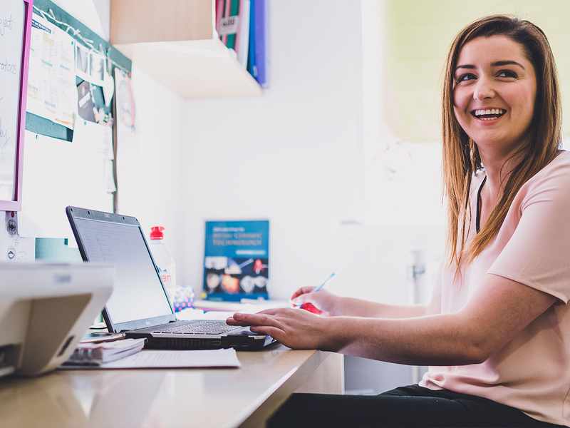 Smiling woman working at her desk with a laptop, taking notes in a bright office space.