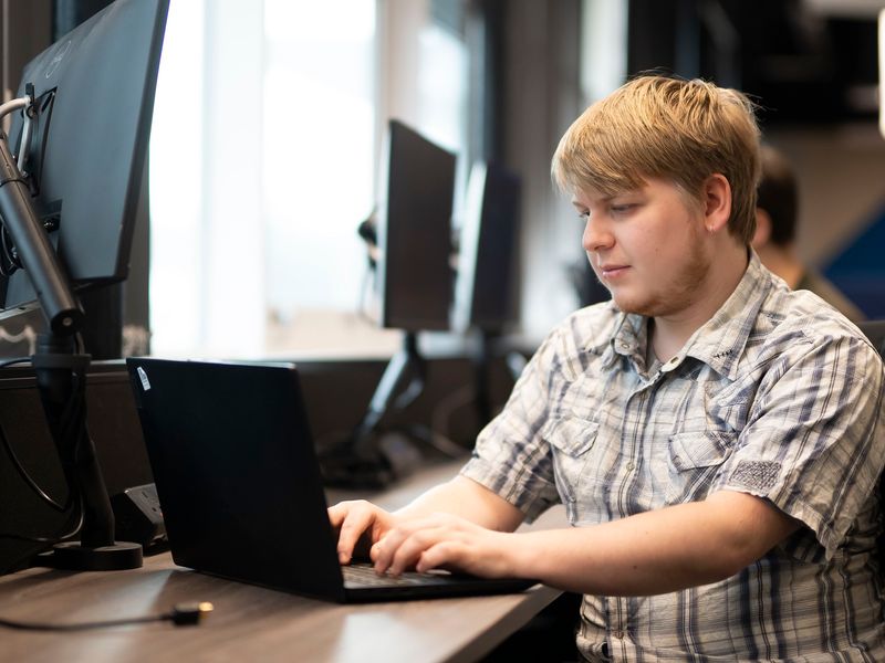 A young man in a short sleeve pattern shirt types on a laptop at a desk