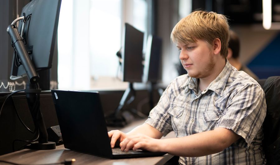 A young man in a short sleeve pattern shirt types on a laptop at a desk
