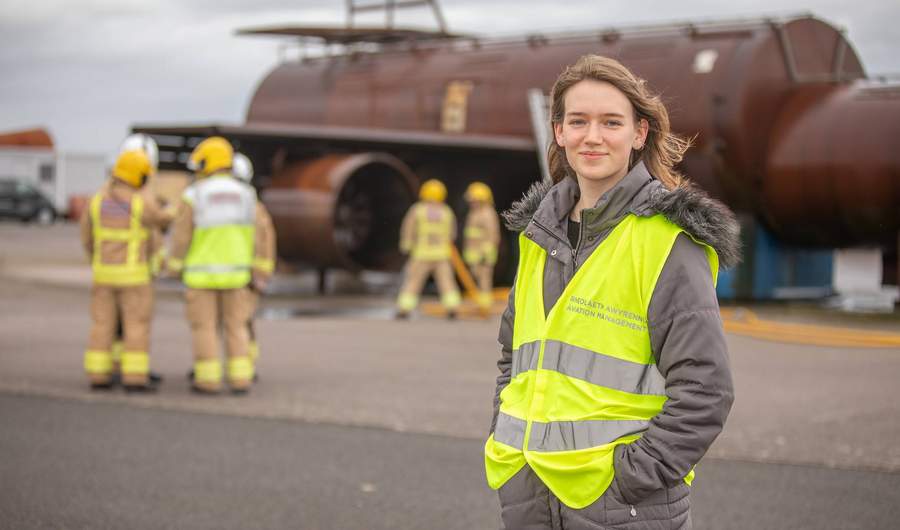 A person wearing a high-vis jacket stands on a tarmacked area. In the background are fire fighters next to a mock-up of an aeroplane fuselage.