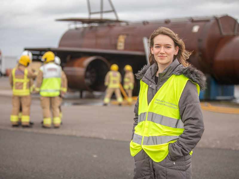 A person wearing a high-vis jacket stands on a tarmacked area. In the background are fire fighters next to a mock-up of an aeroplane fuselage.