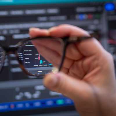 A woman with blonde hair and glasses is seated in front of multiple computer monitors displaying financial data. She appears focused and is touching her nose while working in a modern office setting.