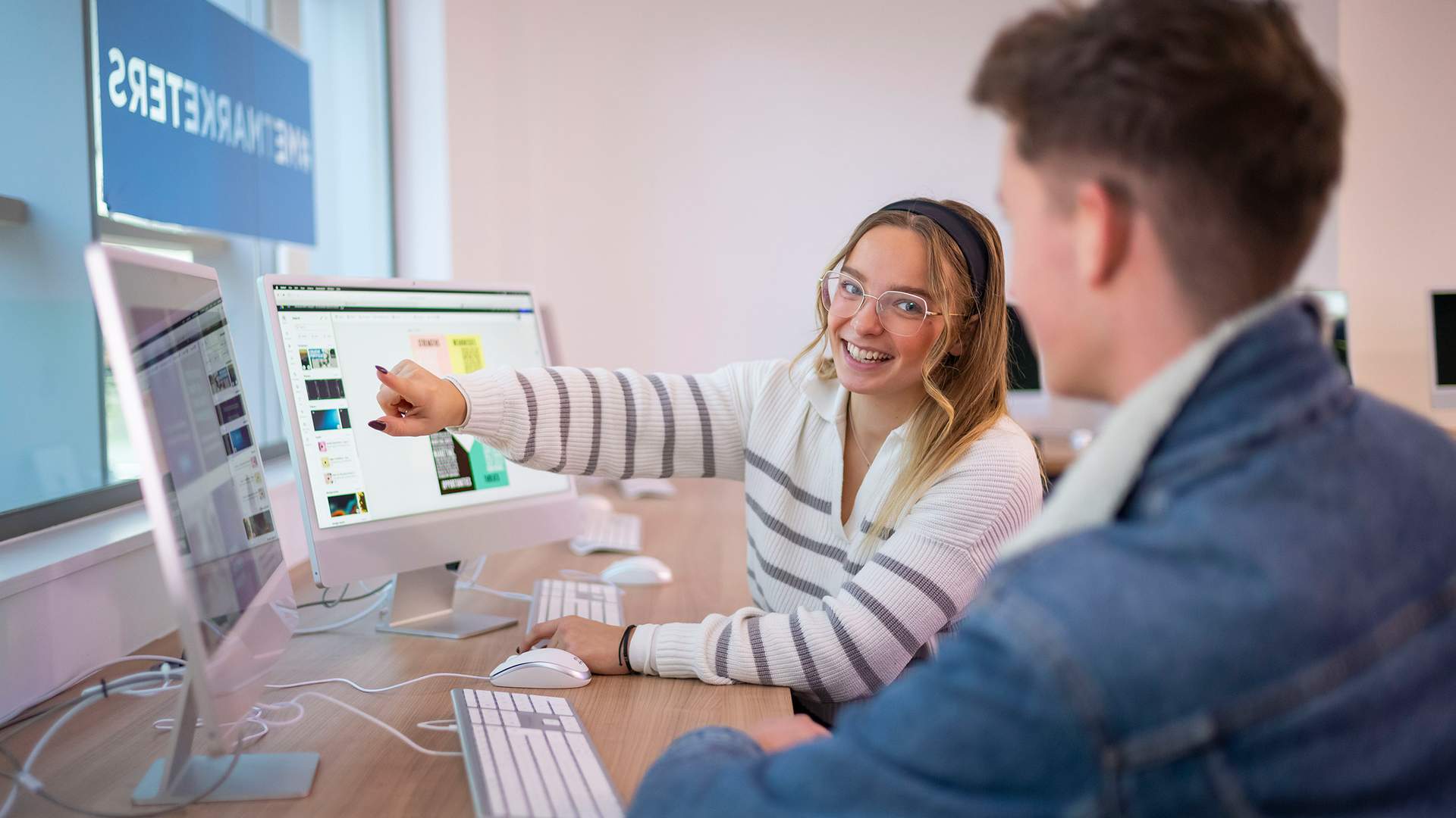 A person with short dark hair and glasses is smiling while sitting outdoors at a table using a laptop. They are wearing a green shirt, and the background is a blurry view of trees with sunlight filtering through.