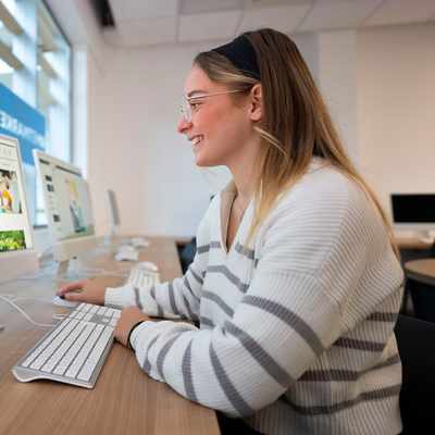 A person with long hair wearing a striped shirt is taking a selfie with a smartphone under a ring light. There are computers and potted plants in the background.