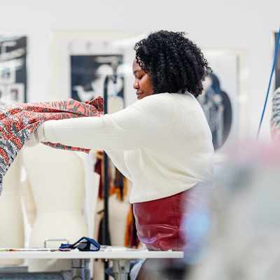 A person with curly hair holds up a colorful fabric in a design studio. In the background, a mannequin and framed art are visible. The room is well-lit, creating an inspiring workspace for creativity.