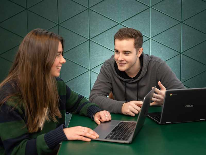 Two individuals seated at a table, each using a laptop, engaged in a collaborative work session.