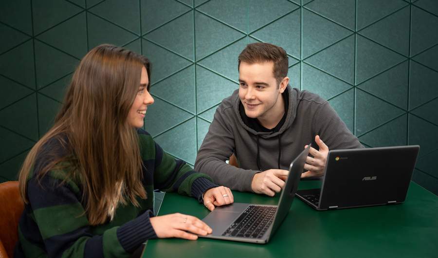 Two individuals seated at a table, each using a laptop, engaged in a collaborative work session.