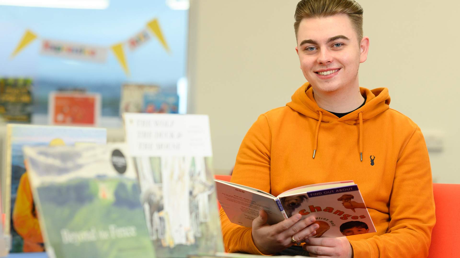 A person in an orange hoodie is smiling while holding a book in a library. There are several books displayed in the foreground. The background shows blurred decor and furniture.