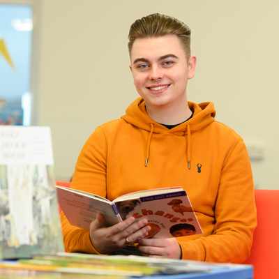 A person in an orange hoodie is smiling while holding a book in a library. There are several books displayed in the foreground. The background shows blurred decor and furniture.