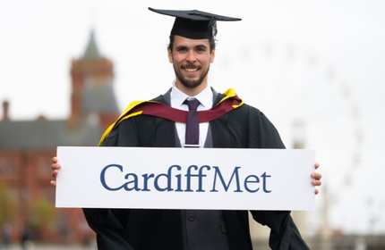 Andrew Fitzsimons, wearing a black graduate cap and gown, holds a Cardiff Met sign in front of their chest