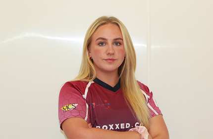 A person with their arms folded stands against a white background. They are wearing a sport team shirt.