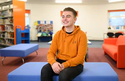 A person wearing an orange hoodie sits smiling on a blue bench in a brightly lit room. The space has colorful furniture, with shelves of books and a bulletin board visible in the background.