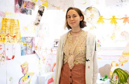 A young woman stands in front of a wall covered with colorful drawings and art. She is wearing a light-colored jacket, a patterned top, and a coral skirt. The art features various abstract and playful designs.