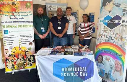 Four individuals stand between a branded Biomedical Science Day display table and posters