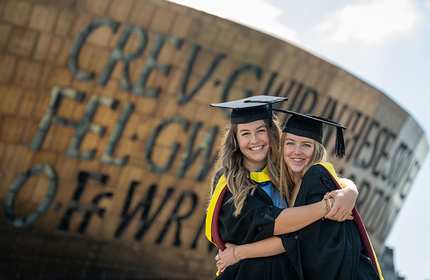 Two graduates in caps and gowns embrace and smile for the camera. They stand in front of a large building with decorative writing. The sky is clear and sunny.