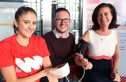 Three people are smiling indoors; a woman on the left in a red shirt is holding a blood pressure monitor, the man in the middle is seated and smiling, and the woman on the right is having her blood pressure checked. Theres a poster in the background.