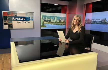 A young woman sits at a news presenter desk holding a piece of paper with both hands