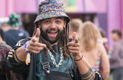 A man with dreadlocks and a colorful bucket hat smiles and points finger guns at the camera. He wears a turquoise patterned shirt, layered necklaces, and stands in a crowd at an outdoor event. The background is pink and blurred.
