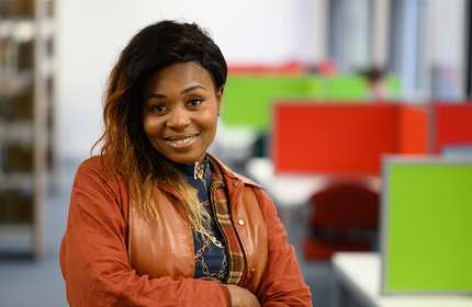 A person with long hair, wearing a brown jacket over a patterned top, smiles while standing in an office with colorful dividers in the background.