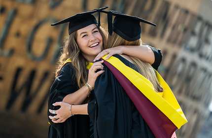 Two female graduates share a heartfelt hug outside a building, symbolizing their friendship and success.