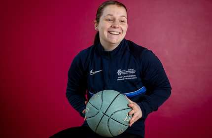 A person smiling and holding a basketball, wearing a dark blue sports jacket, is seated against a burgundy background.