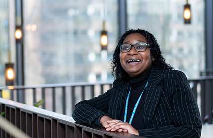 A person wearing a striped suit jacket and glasses is smiling and leaning on a railing. The background features large windows with glowing pendant lights and a blurred cityscape.