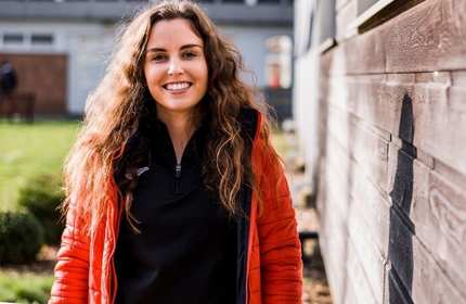 A person with long curly hair smiles while standing outside. They are wearing a red jacket over a black top. The background features a wooden wall and grass, with a softly blurred building in the distance.