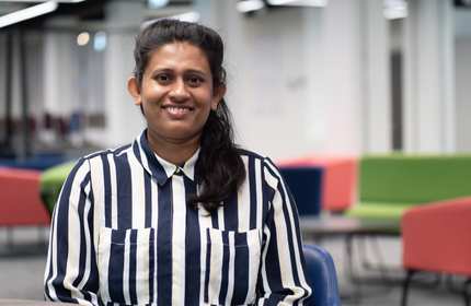 A person with long hair and a striped shirt stands smiling in an open meeting space. There are colourful chairs in the background.