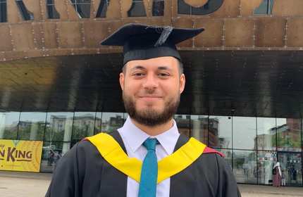Harry Simpson, wearing a graduate cap and gown, stands outsite the Millennium Centre in Cardiff Bay