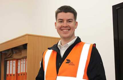 A person stands in an office wearing an orange hi-vis waistcoat.