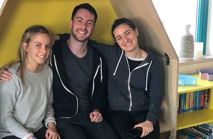 Three people sitting closely in a cozy corner with a yellow background, smiling at the camera. Two women are on either side of a man, each wearing casual clothing. A bookshelf with colorful items is partially visible in the corner.