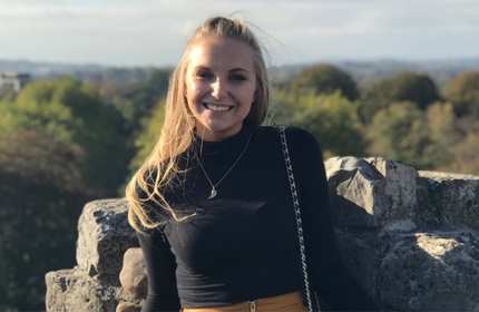 A person with long hair smiles while standing in front of a stone wall. They are wearing a black top and a necklace. The background features a lush landscape with trees under a clear sky.
