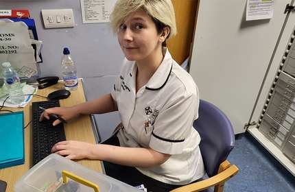 A person with short blond hair is seated at a desk in an office, wearing a white uniform with black trim. They are looking up from a computer keyboard, with files and a water bottle on the desk around them.