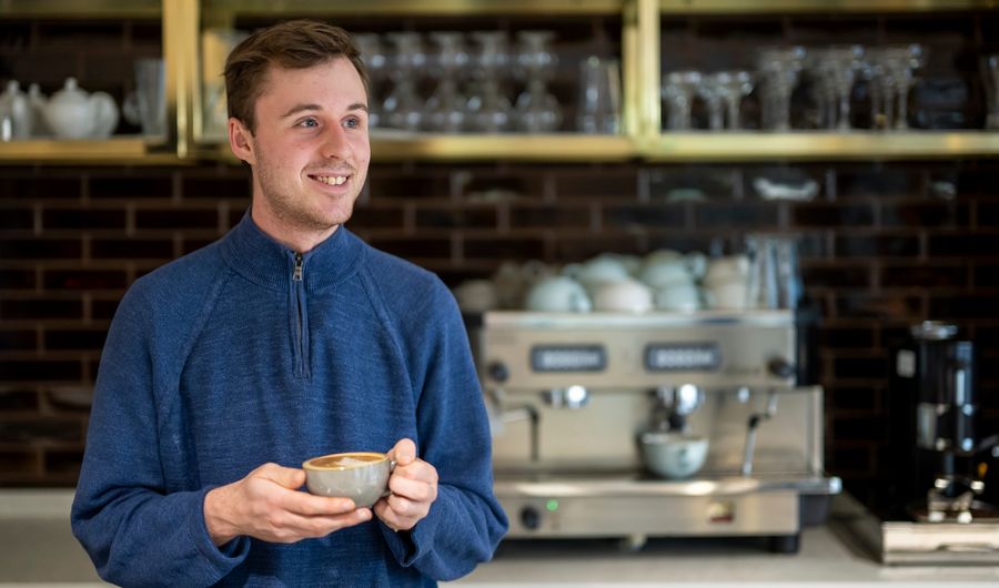 A person holding a coffee cup stands in a bar area. Behind them is a professional coffee machine.