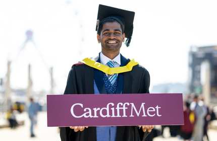 A Cardiff Met student, in graduate cap and gown, holds a purple Cardiff Met sign with both hands