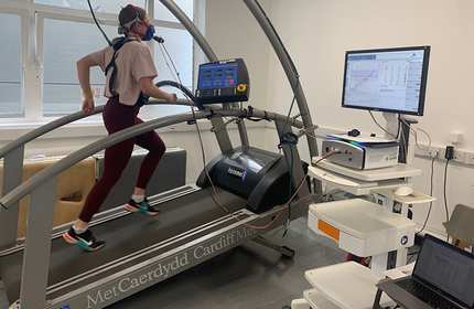 A person is running on a treadmill wearing a mask connected to a machine, likely for a fitness or respiratory test. The treadmill and equipment are inside a lab with monitors displaying data and graphs.