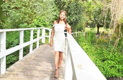 Lerisha, wearing a white dress, stands on a raised wooden walkway above and between trees and shrubbery