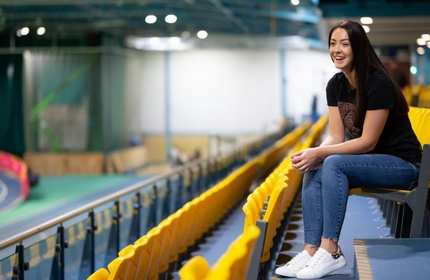 A woman in a black T-shirt and jeans sits smiling on yellow stadium seats overlooking an indoor track. The background includes track lanes and a blue railing. The setting is bright with overhead lighting.