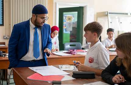 A teacher stands next to a desk talking to a pupil.