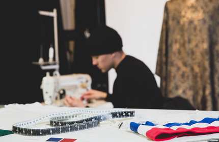 A close-up of a measuring tape and piece of striped fabric. In the background, a person operates a sewing machine.