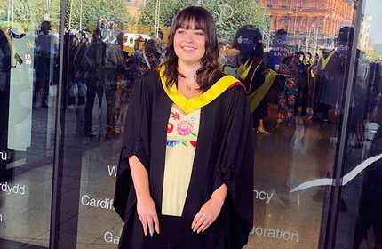 A person in a graduation gown and cap smiles while standing outside a glass-fronted building. They wear a white dress with colorful embroidery underneath their gown. Reflections of people and trees are visible in the glass.