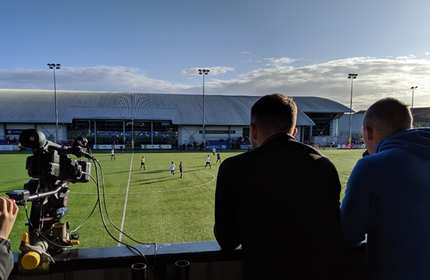 Two mens stand overlooking a game football on an artificial sports pitch at Cardiff Metropolitan University