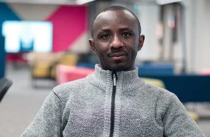 A person wearing a grey cardigan sits smiling in an open meeting space. There are colourful chairs in the background.