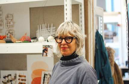 A person with gray hair and glasses stands in a room filled with arts and crafts materials. They are wearing a gray top and smiling. Shelves in the background display various handmade objects and artwork.