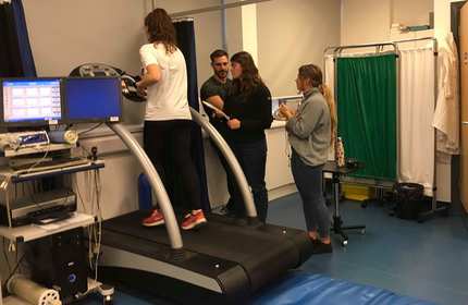 A person walks on a treadmill connected to monitors, while three people observe and take notes in a lab setting. The room has equipment, a curtain, and a blue mat on the floor.