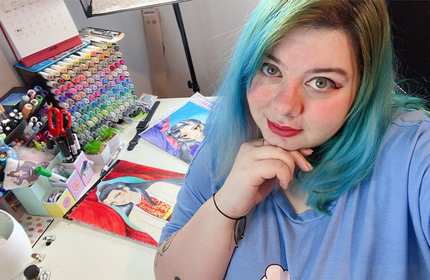 Rachel Stevens takes a selfie while sat next to a desk containing colourful pens and some of their artwork