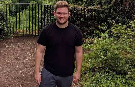 A person with short hair and a beard is standing outdoors on a dirt path, wearing a black t-shirt and gray shorts. The background features lush green foliage and a black metal fence in the distance.