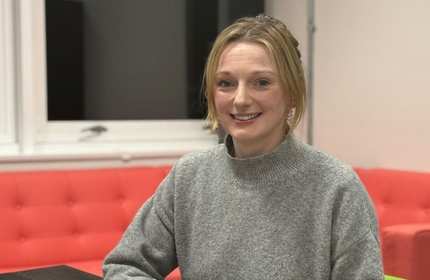 A person sits smiling. Behind them is an orange sofa.