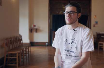 Sam Wilson, wearing a white t-shirt and glasses, sits in a reception room
